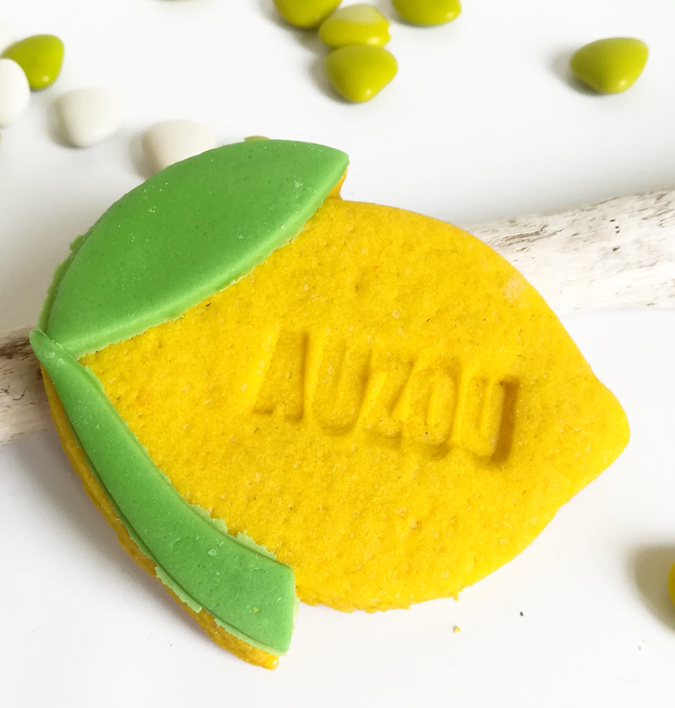 Biscuit sablé en forme de citron. Il est personnalisé avec prénom, marque de la société. Couleur jaune et feuille verte.